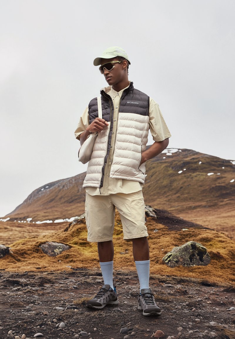 Gilet imbottito nero e crema, camicia beige e pantaloni corti, abbinati a scarpe grigie e calzini azzurri chiari. Il modello si trova su un terreno roccioso.