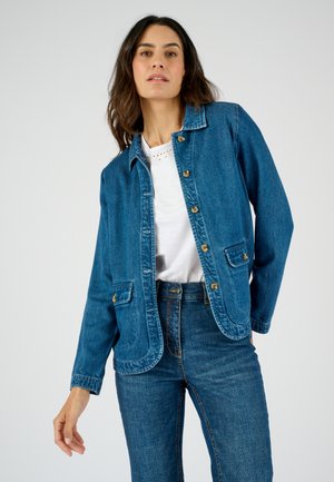 Giacca di jeans di blu medio, con chiusure a bottone, due tasche frontali e orlo curvo. Indossata sopra una maglietta bianca e abbinata a jeans blu.