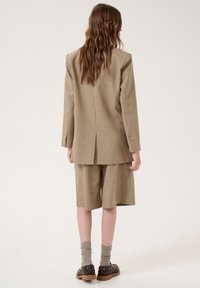 Blaser en lin beige avec des épaules structurées, des poignets boutonnés et un short assorti. Des chaussettes en mélange de laine et des chaussures à plateforme noires complètent le look.