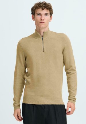 Tan zip-neck sweater made of soft fabric, featuring long sleeves and a textured finish. Side slot detail with ribbed hem. Worn with black pants.