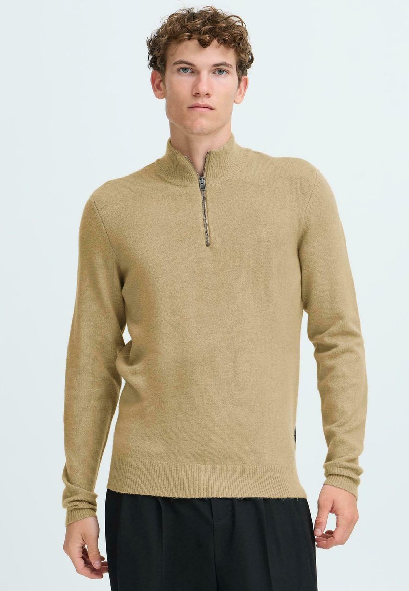 Maglione a collo zip color sabbia, realizzato in tessuto morbido, con maniche lunghe e finitura texturizzata. Dettaglio con fessura laterale e orlo a costine. Indossato con pantaloni neri.