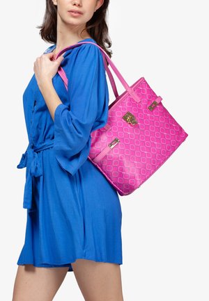 Roze draagtas met geometrisch patroon, glanzende afwerking, dubbele handvatten en details in goudkleurig metaal. Model draagt een blauwe jurk.