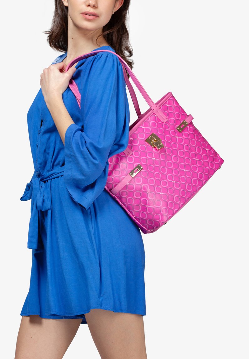 Borsa a tracolla rosa con motivo geometrico, finitura lucida, doppi manici e dettagli in metallo dorato. Il modello indossa un vestito blu.