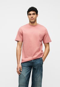 Άνδρας με σγουρά μαλλιά που φοράει ροζ σκονισμένη κοντομάνικη μπλούζα και μπλε τζιν παντελόνι, στέκεται με το ένα χέρι στην τσέπη μπροστά σε λευκό φόντο.