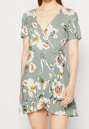 Frau trägt ein kurzärmeliges, salbeigrünes Wickelkleid mit orange-weißem Blumenmuster und Rüschensaum, in der Taille gebunden.