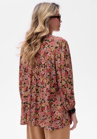 Kobieta z długimi falującymi blond włosami, w okrągłych okularach w tortoise shell, nosi luźną kwiecistą bluzkę w różowe, czerwone i pomarańczowe kwiaty oraz brązowe spodnie.