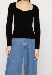 Schwarzes, geripptes Langarmoberteil mit Quadrat-Ausschnitt und taillierter Silhouette, kombiniert mit weit geschnittenen blauen Jeans aus Denim mit verwaschener Textur.