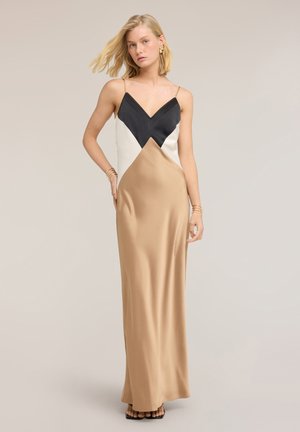 Kobieta w długiej, bezrękawnikowej sukni satynowej z geometrycznymi blokami kolorów czarnych, białych i beżowych, stojąca na jednolitym tle.