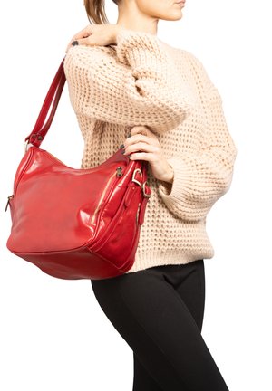 Vrouw in een beige gebreide trui en zwarte broek, die een grote rode leren schoudertas met ritsdetails vasthoudt.