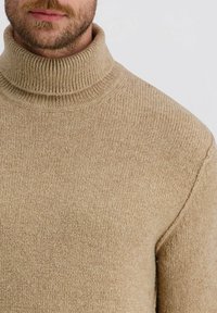 Bärtiger Mann trägt einen beigen, gerippten Rollkragenpullover, der an den Schultern und am Mund gekürzt ist, vor einem schlichten hellen Hintergrund.