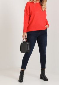 Roter übergroßer Strickpullover, dunkle Skinny Jeans, schwarze Stiefeletten, hält eine kleine schwarze Handtasche mit braunen Griffen.