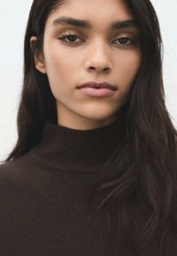 Prenda de cuello alto marrón con textura acanalada, diseño ajustado y cuello alto, llevada por una persona con cabello largo y oscuro y maquillaje neutro.