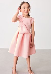 Vestito rosa con un design senza maniche e un grande fiocco decorativo sulla spalla. Tessuto testurizzato con motivi in rilievo. Gonna svasata e lunghezza al ginocchio.