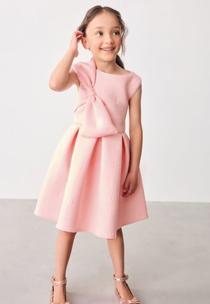 Vestido rosa con un diseño sin mangas y un gran lazo en el hombro. Tejido con textura y patrones en relief. Falda acampanada y hasta la rodilla.
