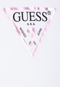 T-shirt blanche ornée d'un logo triangulaire rose et noir avec le texte "GUESS U.S.A" et "WASHED JEANS", accompagné des numéros 1201 et 1203.