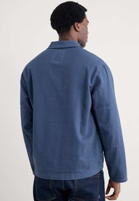 Blå bomullsjacka med krage, långa ärmar och rak nederkant. Har en subtil textur och en rektangulär patch på baksidan.