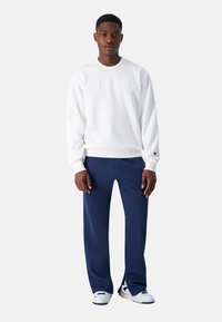 Hvid sweatshirt med præget branding, parret med marineblå bukser. Modellen står i træningssko og viser en afslappet påklædning.