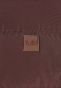Tela burdeos con una textura sutil, que presenta un parche rectangular de cuero marrón grabado con "URBAN CLASSICS" en letras oscuras.