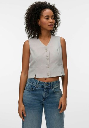 Donna con capelli ricci che indossa un gilet beige senza maniche con bottoni e jeans blu a vita alta, guarda leggermente verso destra su uno sfondo semplice.