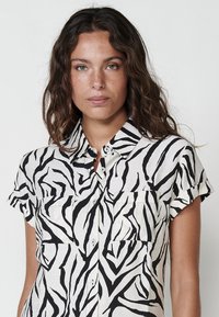 Camisa de botones de manga corta con estampado de cebra, que presenta una paleta de colores en blanco y negro, dos bolsillos en el pecho y un cuello. 