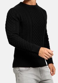 Zwarte gebreide trui met een gestructureerd kabelpatroon, voorzien van een ronde hals en geribde mouwen. Het materiaal lijkt dik en warm.