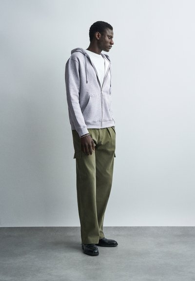 Άνδρας που στέκεται και κοιτά προς τα πλάγια, φοράει γκρι φούτερ με φερμουάρ, λευκό πουκάμισο, χακί παντελόνι cargo και μαύρα παπούτσια σε γκρι πάτωμα.