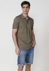 Polo en verde oliva texturizado con una tapeta negra de botones, combinado con pantalones cortos de mezclilla azules con dobladillos enrollados.