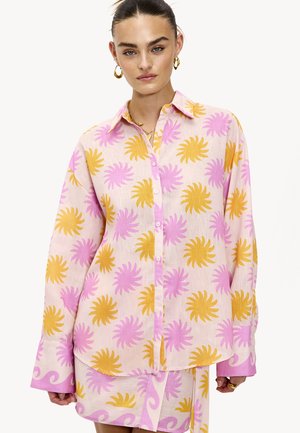Chemise à manches longues en tissu léger, présentant un motif de feuilles roses et jaunes sur un fond rose clair. Design à boutons sur le devant avec un col.