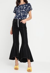 Námořnický květinový top s krátkými rukávy, kombinovaný s černými rozšířenými kalhotami s bílým prošíváním a detailem na zip, doplněný žlutými sandály.