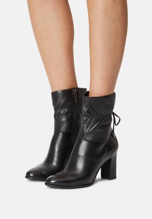 Svarta läderstövlar till mitten av vaden med klackar i blockstil, sidodragkedjor och samlade detaljer, barfota mot en vit bakgrund.