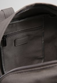 Vnitřek šedého textilního batohu s kapsou na zip a štítkem. Stěny mají strukturované tkaní, což zajišťuje odolnost a organizaci.