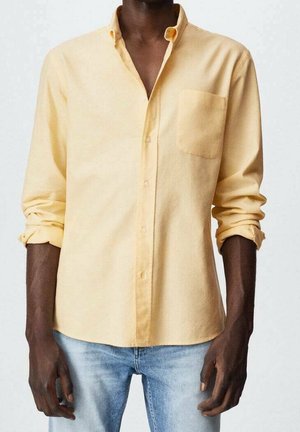 Homme portant une chemise à boutons jaune clair avec les manches retroussées et un jean bleu clair, sur un fond uni.