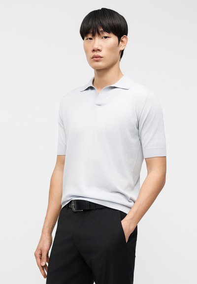 Ανοιχτό γκρι κοντομάνικο πόλο μπλουζάκι με κλασικό γιακά και ομαλή υφή, φορεμένο με μαύρο παντελόνι και ζώνη, στέκεται μπροστά σε ουδέτερο φόντο.