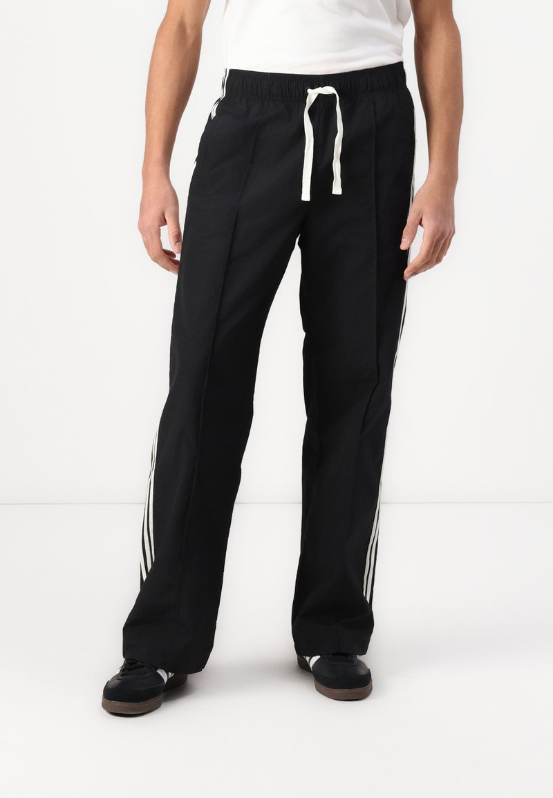 Black athletic pants with a drawstring waist, featuring white side stripes and a lightweight fabric. Straight-leg design with pockets.