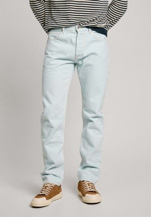 Jeans in denim leggermente blu con taglio dritto, dotati di cinque tasche e dettagli di cucitura sottili, abbinati a sneakers marroni e crema.