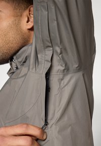 Ανοιχτό γκρι μπουφάν με κοντινή λήψη ενός τσεπιδίου με φερμουάρ, που παρουσιάζει λεία υφή και κομψό σχεδιασμό, υπογραμμίζοντας την ανθεκτικότητα του υφάσματος.