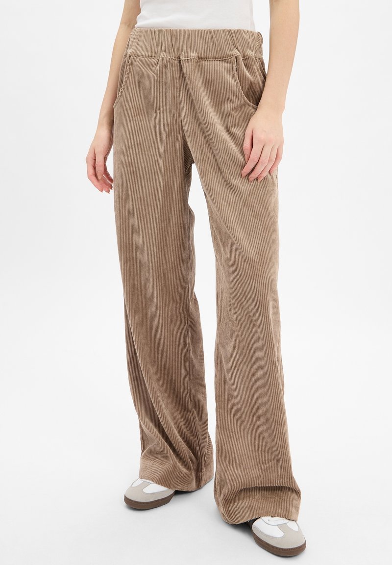 Femme portant un pantalon large en velours côtelé beige avec taille élastique et baskets blanches, debout devant un fond clair uni.