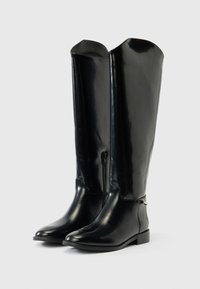 Ζευγάρι από ψηλές, μαύρες, γυαλιστερές δερμάτινες μπότες με χαμηλά τακούνια και φερμουάρ στο πλάι, όρθιες πάνω σε λευκή επιφάνεια.