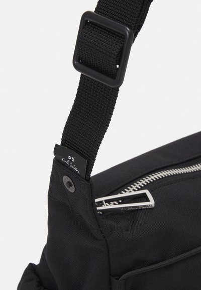 Borsa in tela nera con tracolla e fibbia rettangolare nera, dotata di una cerniera argentata e un'etichetta con logo. Tessuto testurizzato con dettagli di cucitura visibili.
