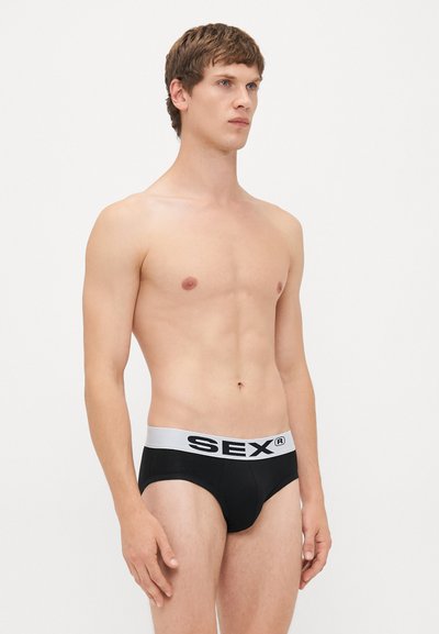 Junger Mann mit kurzen braunen Haaren, der schwarze Unterhose mit einem weißen Bund trägt, auf dem fettgedruckte schwarze Buchstaben stehen, und vor einem schlichten weißen Hintergrund steht.