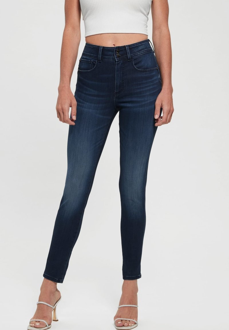Dunkelblaue Skinny-Jeans mit hoher Taille, subtilen Wischdetails und klassischem Fünf-Taschen-Design, kombiniert mit eleganten Sandalen.