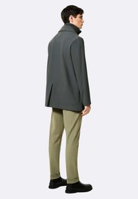 Cappotto grigio con colletto alto, spacco posteriore, abbinato a pantaloni verde oliva chiaro e calzature nere, visto da un lato posteriore.