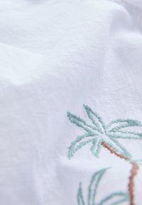 Tissu blanc texturé avec un motif de palmier brodé en vert et marron. Matériau doux et léger avec un léger éclat.