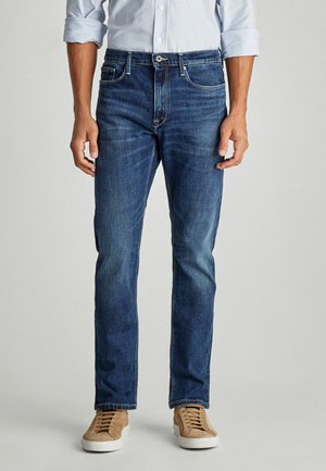 Jeans de denim azul oscuro con un corte recto, que presentan un diseño clásico de cinco bolsillos y costuras en contraste, combinados con zapatillas de ante color marrón.