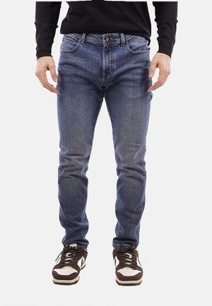 Jeans slim fit blu scuro con una texture scolorita, cinque tasche e chiusura con bottone, abbinati a sneakers marroni e bianche.