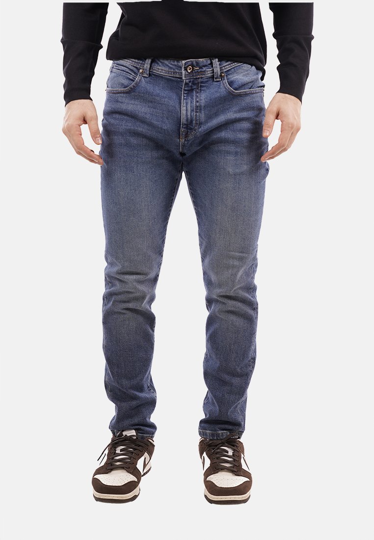 Jeans slim fit blu scuro con una texture scolorita, cinque tasche e chiusura con bottone, abbinati a sneakers marroni e bianche.