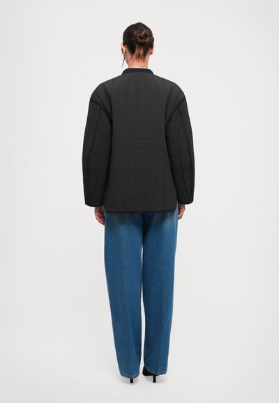 Eine Frau steht mit dem Rücken zur Kamera, trägt eine schwarze Steppjacke, blaue weite Jeans und schwarze High Heels vor einem schlichten hellen Hintergrund.
