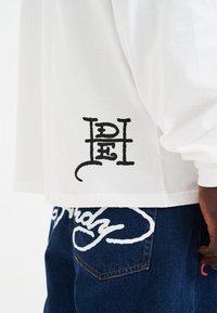 Weiße Langarmbluse mit einem schwarzen, gestickten Logo am Saum, kombiniert mit blauen Denim-Jeans, die weiße, geschwungene Akzente aufweisen.