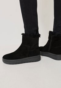 Fekete velúr bokacsizmák műszőrme díszítéssel a tetején és oldalcipzárral, fekete gumi talppal és texturált részletekkel.