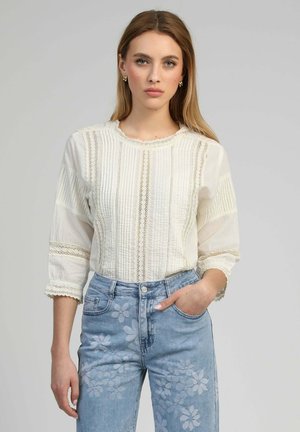 Jeune femme portant un chemisier en dentelle blanche et un jean bleu clair avec des motifs floraux, debout devant un fond gris uni.